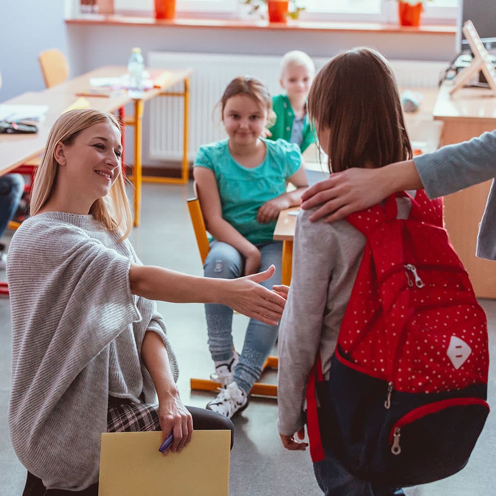 Female teacher shaking a young girl's hand as her parent drops her off at school