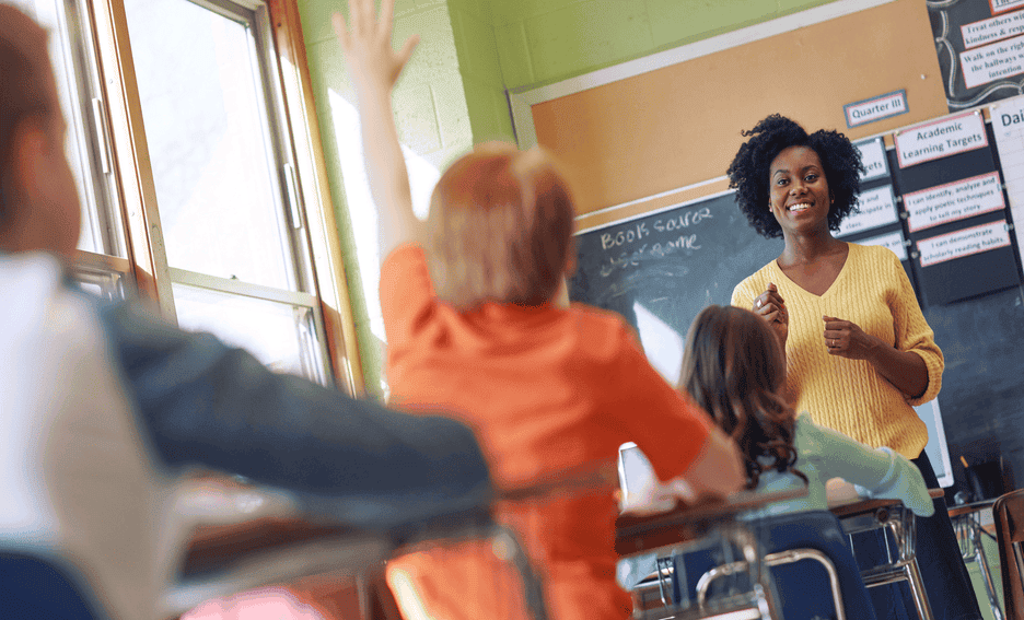 Student raising his hand while a teacher explains a lesson in a classroom