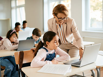 Happy teacher assists schoolgirl in using laptop during computer class in the classroom